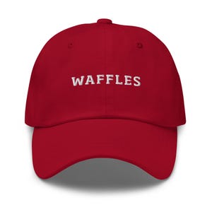 May include: A red baseball cap with the word "WAFFLES" embroidered in white across the front. The cap has a curved brim and a rounded crown. This hat is a casual accessory.
