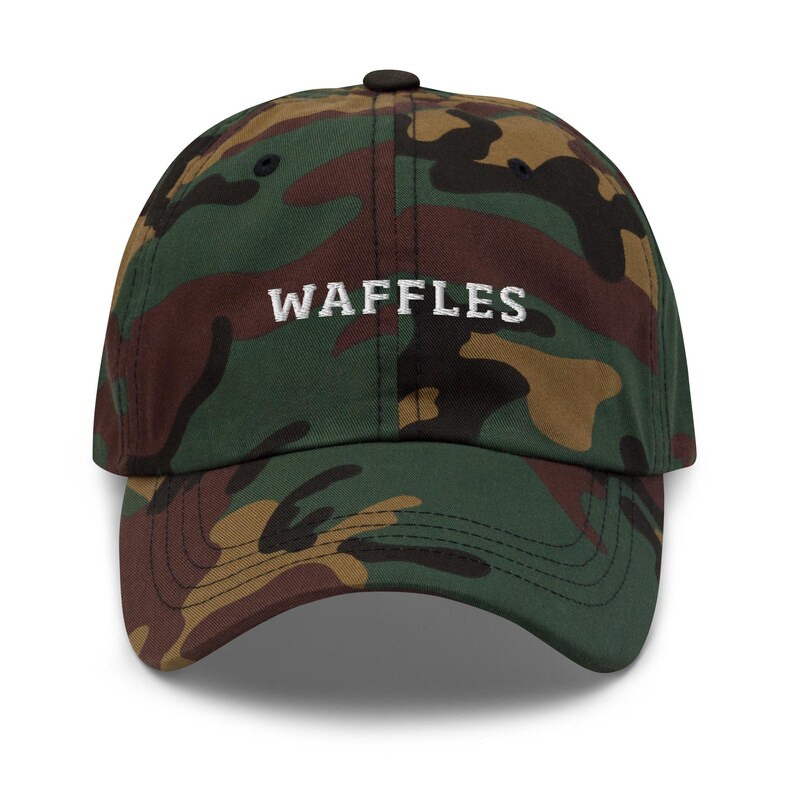 May include: A camouflage baseball cap with the word "WAFFLES" embroidered in white across the front. The cap features a curved brim and a black button on top. The camouflage pattern includes shades of green, brown, and tan.