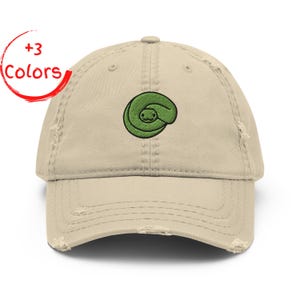 Può includere: Un cappellino da baseball beige consumato con un design a serpente verde ricamato. Il cappellino ha una visiera curva e un aspetto vintage usurato. Il testo "+3 Colors" è in un cerchio rosso.