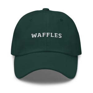 May include: A dark green baseball cap with the word "WAFFLES" embroidered in white across the front. The cap has a curved brim and a button on top. This hat is a casual accessory.