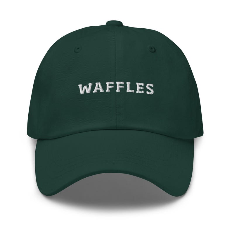 May include: A dark green baseball cap with the word "WAFFLES" embroidered in white across the front. The cap has a curved brim and a button on top. This hat is a casual accessory.