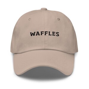 May include: A beige baseball cap with the word "WAFFLES" embroidered in black across the front. The cap has a curved brim and an adjustable strap at the back. The hat is made of a soft, durable fabric.