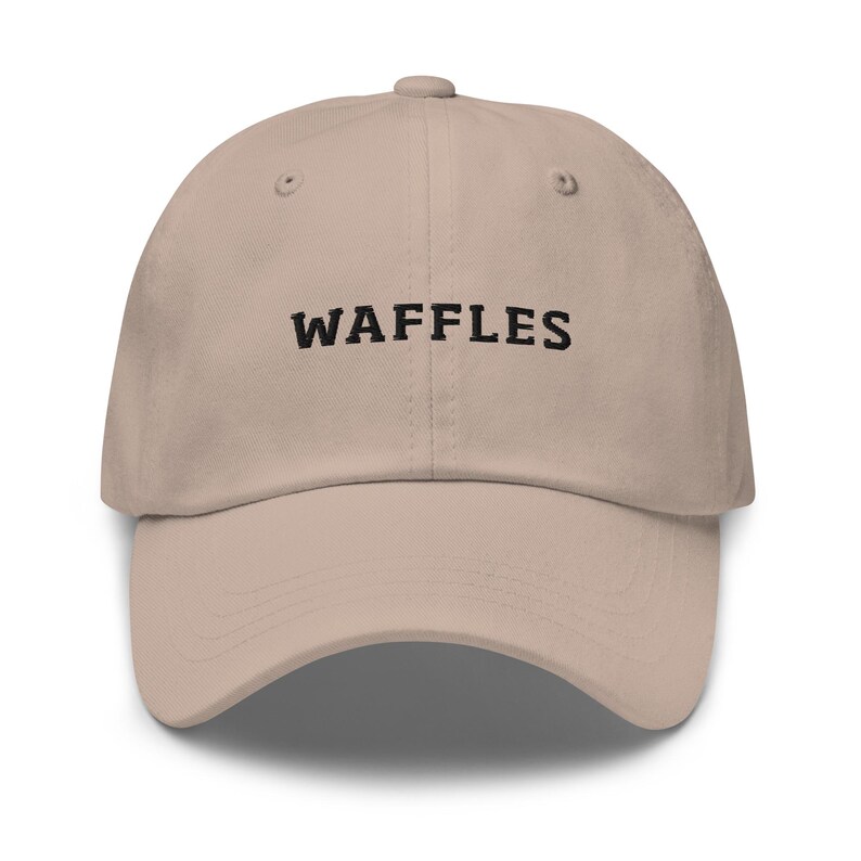 May include: A beige baseball cap with the word "WAFFLES" embroidered in black across the front. The cap has a curved brim and an adjustable strap at the back. The hat is made of a soft, durable fabric.