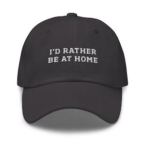 May include: A dark gray baseball cap with the embroidered text "I'D RATHER BE AT HOME" in white. The cap has a curved brim and a structured fit, suitable for casual wear.