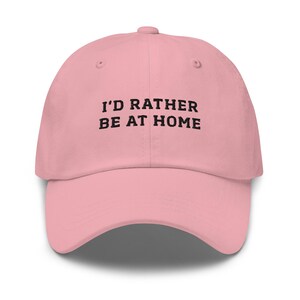 May include: A light pink baseball cap with the text "I'D RATHER BE AT HOME" in black block letters. The cap has a curved brim and a rounded crown. The hat is made of a soft material.