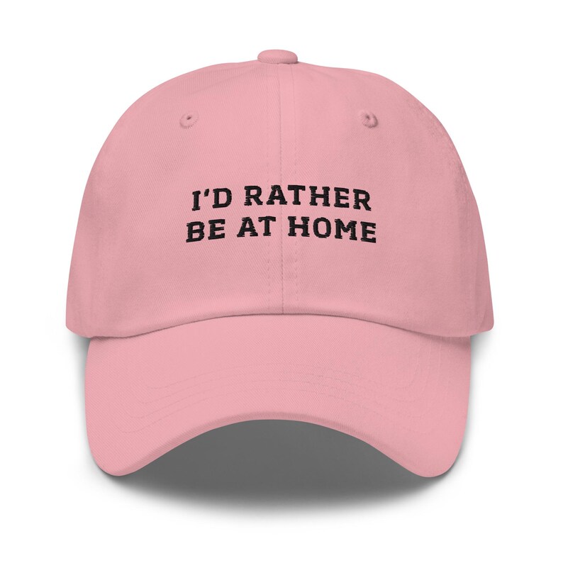 May include: A light pink baseball cap with the text "I'D RATHER BE AT HOME" in black block letters. The cap has a curved brim and a rounded crown. The hat is made of a soft material.