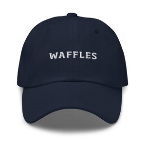 May include: A navy blue baseball cap with the word "WAFFLES" embroidered in white across the front. The cap has a curved brim and a button on top. This hat is a casual accessory.