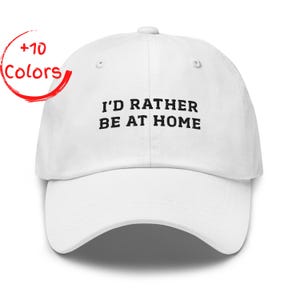 May include: White baseball cap with the embroidered text "I'D RATHER BE AT HOME" in black. The cap has a curved brim and a button on top. A red circle with the words "+10 Colors" is in the upper left corner.