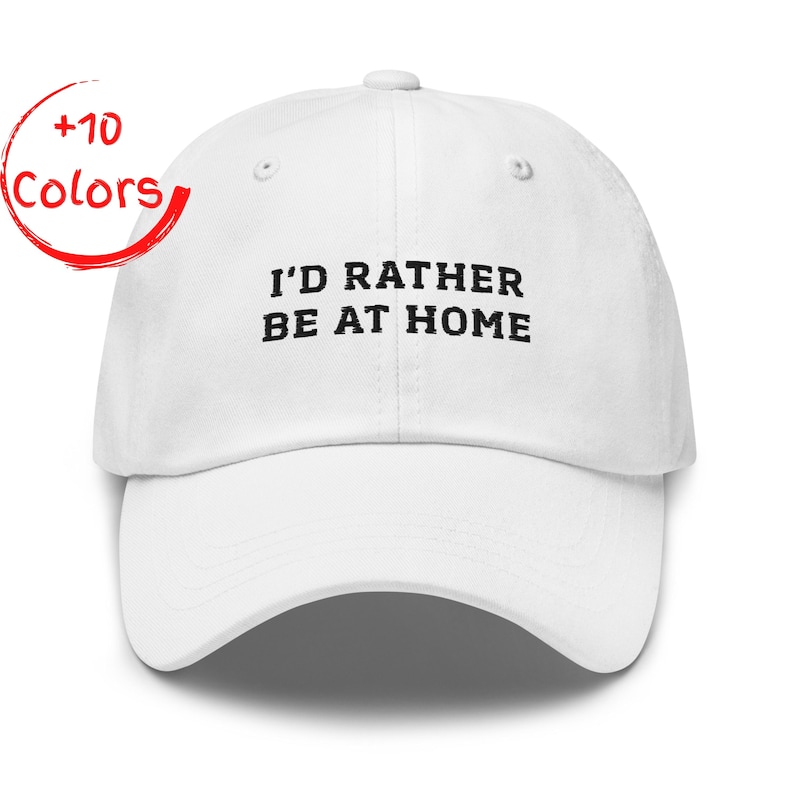 May include: White baseball cap with the embroidered text "I'D RATHER BE AT HOME" in black. The cap has a curved brim and a button on top. A red circle with the words "+10 Colors" is in the upper left corner.