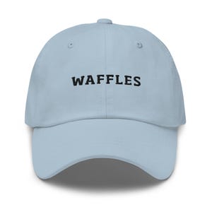 May include: A light blue baseball cap with the word "WAFFLES" embroidered in black across the front. The cap has a curved brim and an adjustable strap at the back. The hat is made of a soft, durable material.