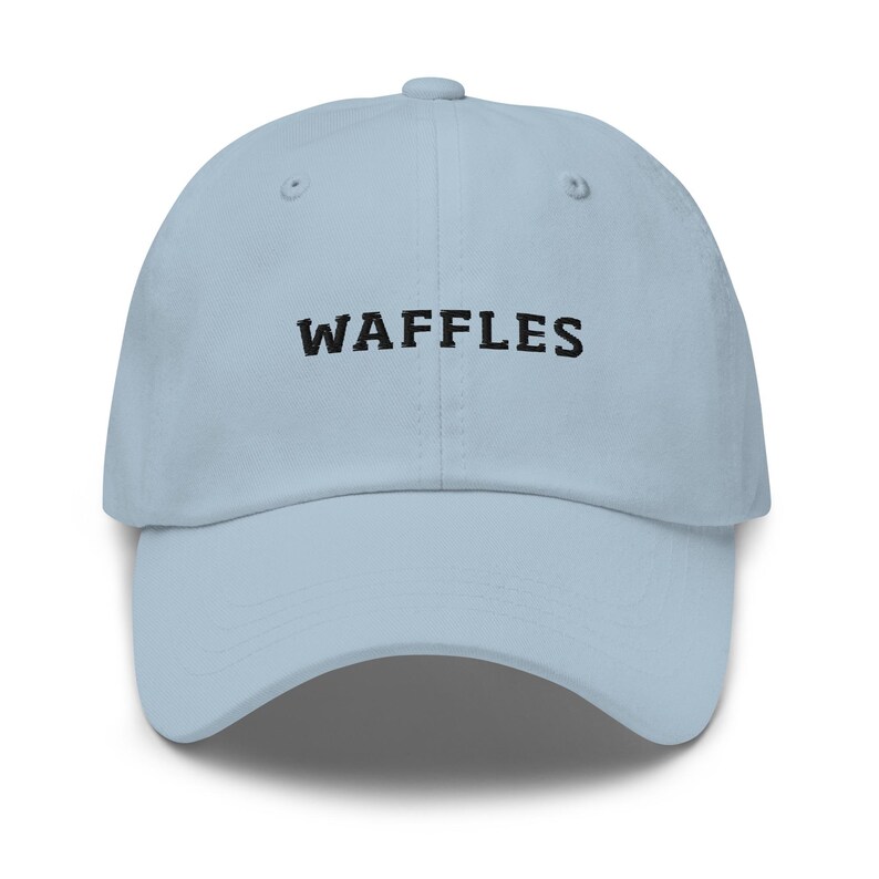 May include: A light blue baseball cap with the word "WAFFLES" embroidered in black across the front. The cap has a curved brim and an adjustable strap at the back. The hat is made of a soft, durable material.
