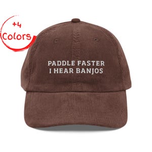 May include: A brown corduroy baseball cap with the embroidered text "PADDLE FASTER I HEAR BANJOS". The cap has a curved peak and a button on top. A red circle with "+4 Colors" is in the upper left corner.
