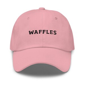 May include: A light pink baseball cap with the word "WAFFLES" embroidered in black across the front. The cap has a curved brim and a button on top. This hat is a casual accessory.