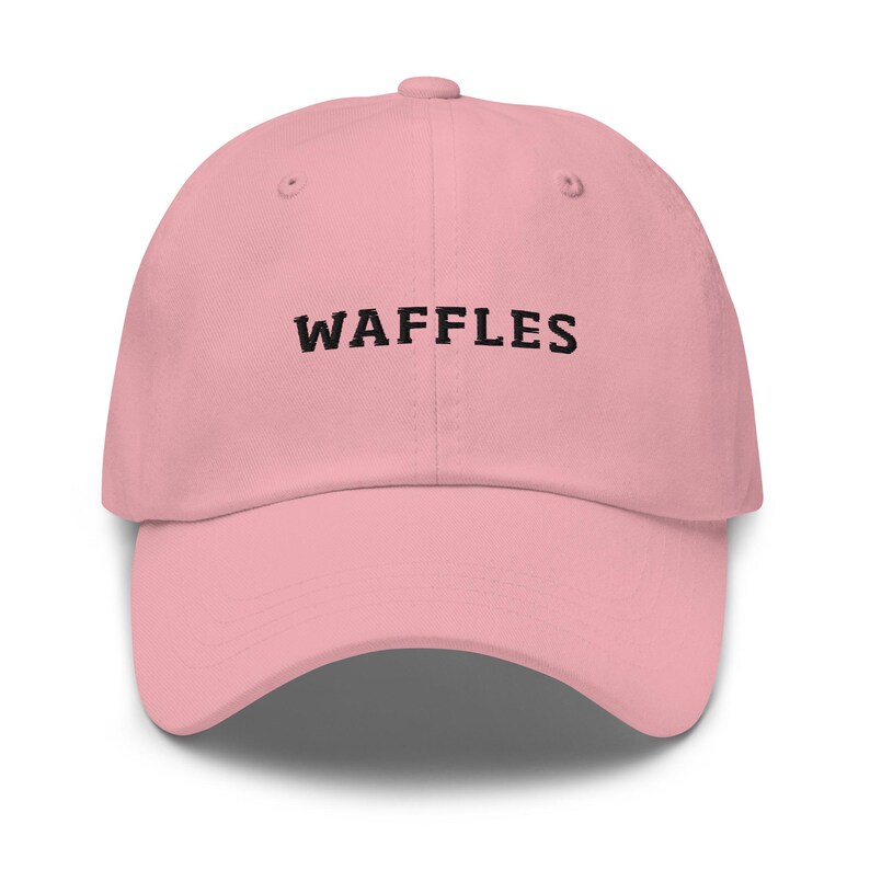May include: A light pink baseball cap with the word "WAFFLES" embroidered in black across the front. The cap has a curved brim and a button on top. This hat is a casual accessory.