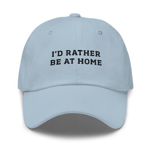 May include: A light blue baseball cap with the text "I'D RATHER BE AT HOME" in black block letters. The cap has a curved brim and a six-panel design.