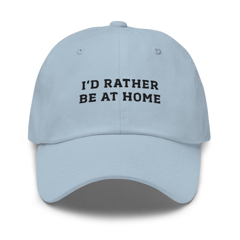 May include: A light blue baseball cap with the text "I'D RATHER BE AT HOME" in black block letters. The cap has a curved brim and a six-panel design.