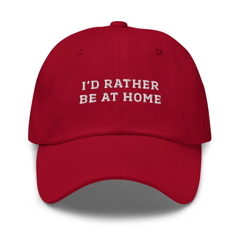 May include: A red baseball cap with the embroidered text "I'D RATHER BE AT HOME" in white. The cap has a curved brim and a six-panel design, suitable for casual wear.