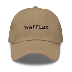 May include: A tan baseball cap with the word "WAFFLES" embroidered in black across the front. The cap has a curved brim and a button on top.