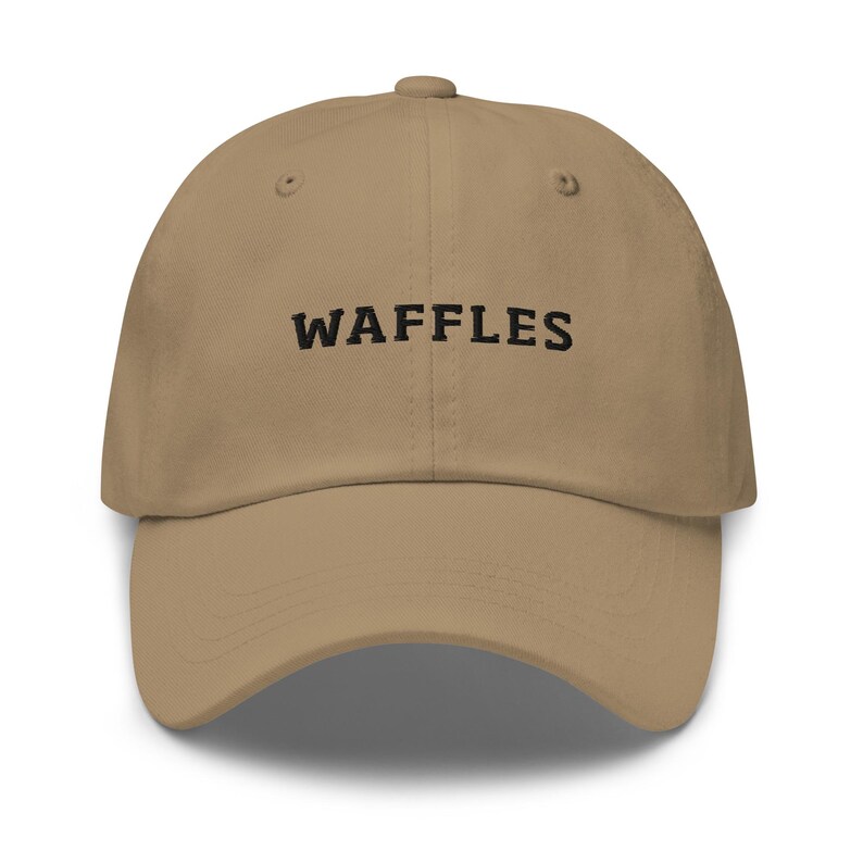 May include: A tan baseball cap with the word "WAFFLES" embroidered in black across the front. The cap has a curved brim and a button on top.