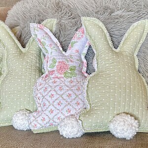 May include: Three decorative bunny-shaped pillows. Two are light green with white polka dots, and one is white with a pink floral pattern. Each has a fluffy white tail and a scalloped edge. The pillows are made of quilted fabric.