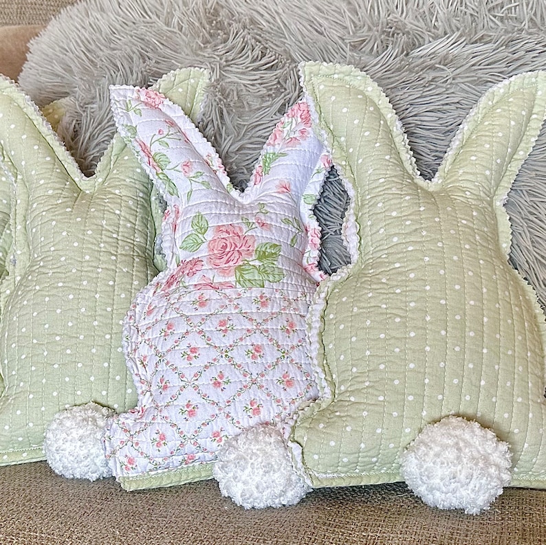 May include: Three decorative bunny-shaped pillows. Two are light green with white polka dots, and one is white with a pink floral pattern. Each has a fluffy white tail and a scalloped edge. The pillows are made of quilted fabric.