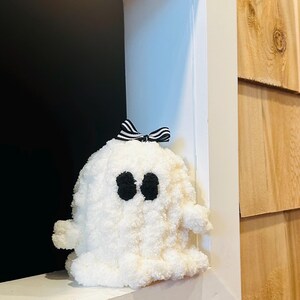 May include: A fluffy white ghost-shaped plush toy with black eyes and a black and white striped bow. The plush is sitting in a white window frame, with a dark background and wood siding.