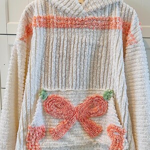 May include: Cream-colored hooded sweatshirt with a textured, corduroy-like design. Features a coral-colored horizontal stripe across the chest and a large coral bow on the front pocket. The cuffs and pocket corners are also coral.
