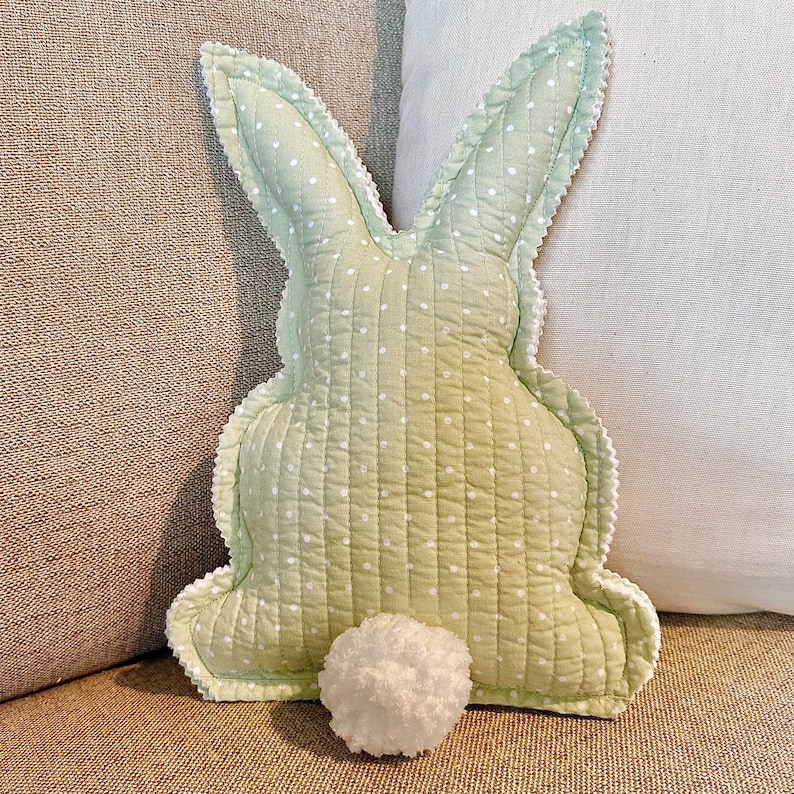 May include: A light green bunny-shaped pillow with white polka dots and a white pom-pom tail. The pillow has a quilted texture and a scalloped edge. The pillow is a decorative item.