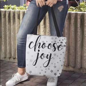 May include: A white tote bag featuring a repeating pattern of small gray stars and the black cursive text "choose joy" centered on the front. The bag has two black shoulder straps and a rectangular shape, constructed from a fabric material.