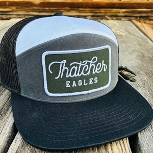 May include: A trucker cap with a black peak, a white crown, and a grey central panel. The cap has a green patch with the words "Thatcher Eagles" in white script.