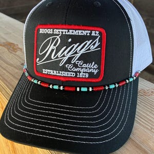 Puede incluir: Gorra de camionero negra y blanca con la parte trasera de malla blanca. La parte delantera presenta un parche con el texto "Riggs Cattle Company" y "Established 1879". Una banda de cuentas rojas, blancas y turquesas adorna la visera.