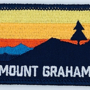 May include: Embroidered patch featuring a mountain landscape with a sunset design. The patch has a navy blue border and a colorful sky with yellow, orange, and red hues. The text "MOUNT GRAHAM" is embroidered in white.