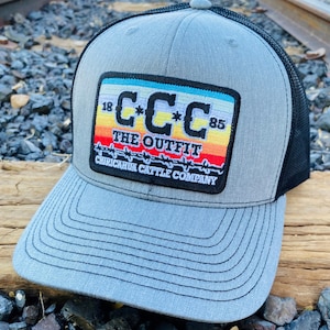 May include: A gray and black trucker hat with a patch that reads "CCC The Outfit Chiricahua Cattle Company" with a rainbow stripe design and barbed wire.