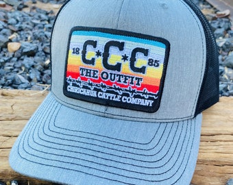 May include: A gray and black trucker hat with a patch that reads "CCC The Outfit Chiricahua Cattle Company" with a rainbow stripe design and barbed wire.