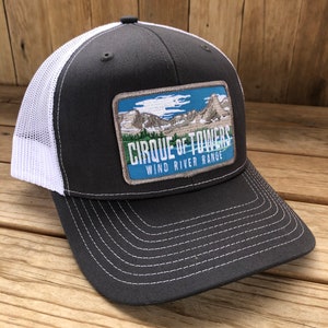 May include: A dark gray and white trucker hat with a patch that reads "Cirque of Towers Wind River Range". The patch features a blue sky with white clouds and a mountain range.