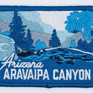 May include: An embroidered patch with a blue background featuring a scenic landscape of a river flowing through a canyon with trees and mountains. The text "Arizona Aravaipa Canyon" is embroidered in white on the bottom of the patch.
