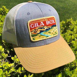 May include: A blue and white trucker hat with a tan brim. The hat features a patch with the text "Arizona Gila Box" and a scenic image of a river.