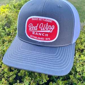 Pode incluir: Boné de caminhoneiro cinza e branco com um patch oval vermelho que diz "Riggs Settlement Red Wing Ranch Established 1879".