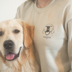 Pu&ograve; includere: Una felpa beige con un'illustrazione in bianco e nero di una testa di cane e il nome "Waylon" stampato sul davanti. Un golden retriever &egrave; seduto accanto alla persona che indossa la felpa.