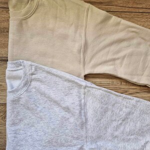 Pu&ograve; includere: Due felpe con maniche lunghe, una di colore beige chiaro e l'altra di colore grigio melange. Entrambe le felpe sono piegate e disposte su una superficie di legno.