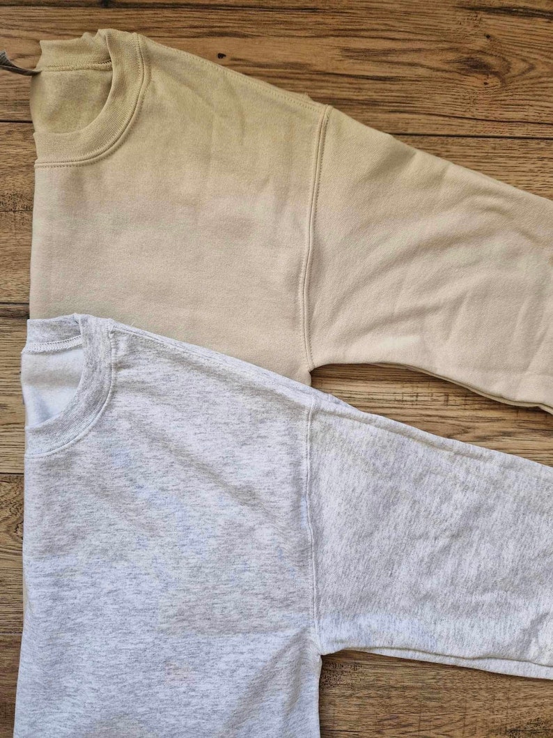 Pu&ograve; includere: Due felpe con maniche lunghe, una di colore beige chiaro e l'altra di colore grigio melange. Entrambe le felpe sono piegate e disposte su una superficie di legno.