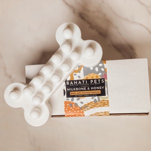 Op de afbeelding: Wit kauwspeeltje voor honden in de vorm van een bot met verhoogde bulten. Het speeltje ligt naast een doos met een label waarop staat: "BAHATI PETS FUR CLEANSER FOR DOGS MILKBONE & HONEY Made with Colloidal Oatmeal NET WEIGHT 1 (OZ) 143 GRAMS"