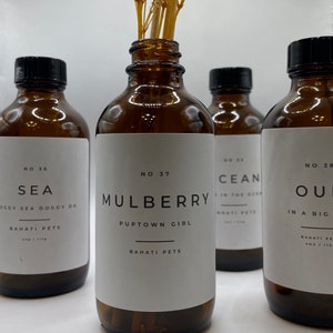 May include: Four amber glass bottles with white labels and black caps. The labels have the text "NO 36 SEA", "NO 37 MULBERRY PUPTOWN GIRL", "NO 33 CEAN IN THE OCEAN", and "NO 38 OU IN A BIG". Each bottle has the text "BAHATI PETS" on the label. A bouquet of dried white flowers is in the center of the image.