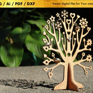 Op de afbeelding: Een houten ringhouder in de vorm van een boom met ingewikkelde details, waaronder harten, sterren en bloemen. De houder is gemaakt van lichtbruin hout en heeft een natuurlijke afwerking. Hij staat op een grijs oppervlak.