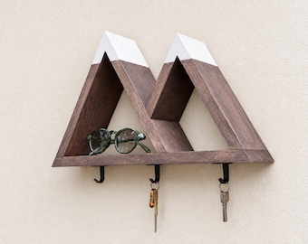 May include: A wall-mounted wooden shelf shaped like two mountain peaks with dark brown stained wood and white painted tops. The shelf features three black metal hooks underneath, with two metal keys hanging from the hooks and a pair of glasses.