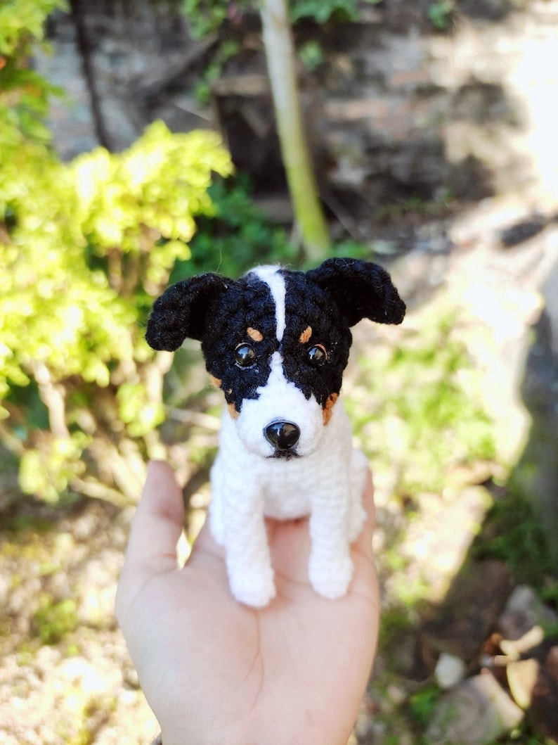 May include: A crocheted stuffed dog toy with black and white fur and brown eyes. The dog is sitting on a person's hand.