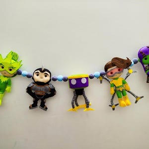 May include: A felt banner featuring five superhero-themed figures in vibrant colors. The figures include a green-clad character with a leaf-like headpiece, a black and gray armored figure, a purple and yellow robot, a yellow and green character with goggles, and a purple-clad character.