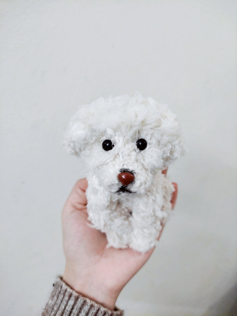May include: A white fluffy toy dog with black eyes and a brown nose. The dog is being held in a person's hand.
