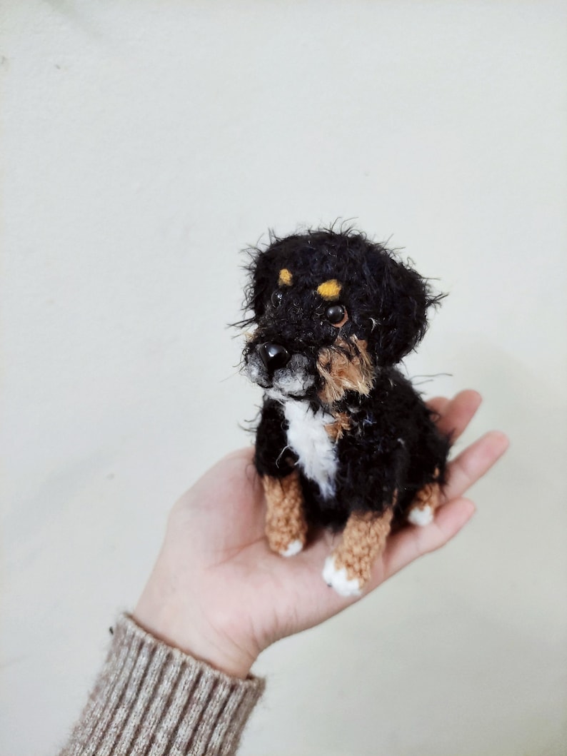 May include: A small crocheted Bernese Mountain Dog puppy toy with black, brown, and white fur. The toy is sitting in the palm of a hand.