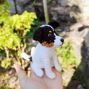 May include: A crocheted dog toy in white, black, and brown. The dog is sitting up with its head tilted to the side. The toy is sitting on a person's hand.